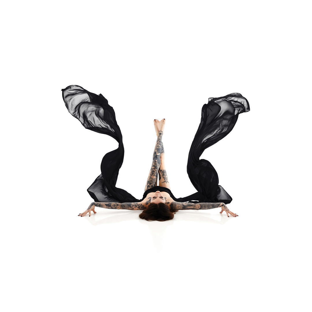 Foto artística en clave alta de una mujer tatuada tumbada de espaldas con las piernas grácilmente levantadas y una tela negra transparente que fluye de sus brazos, símbolo de empoderamiento y autoexpresión en la colección Self-Love de Nuovo.
