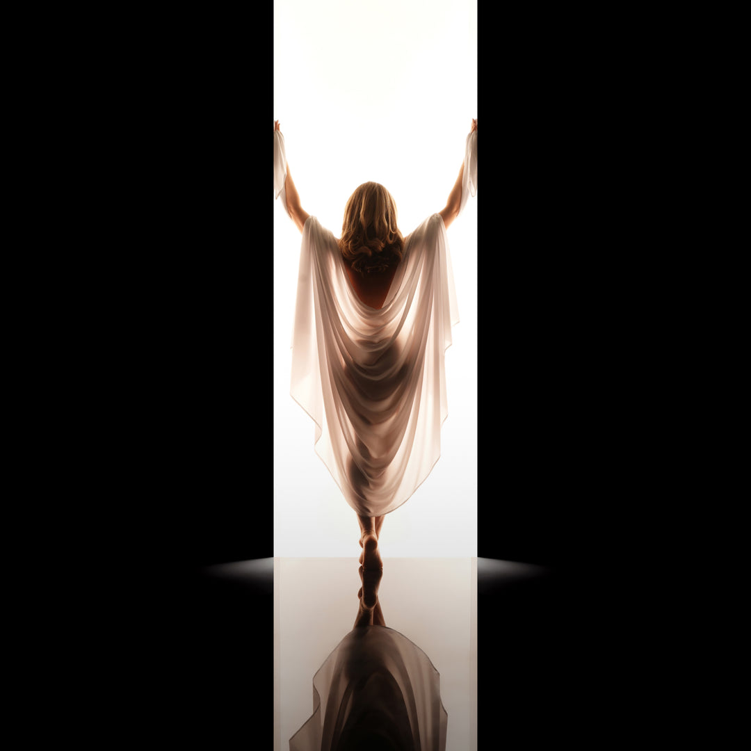 Woman draped in flowing cream fabric poses from behind in a backlit studio doorway for an elegant fine art boudoir portrait.