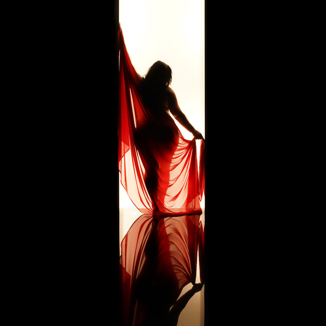 Backlit fine art silhouette of a woman draped in sheer red fabric in a doorway of light, with floor reflection