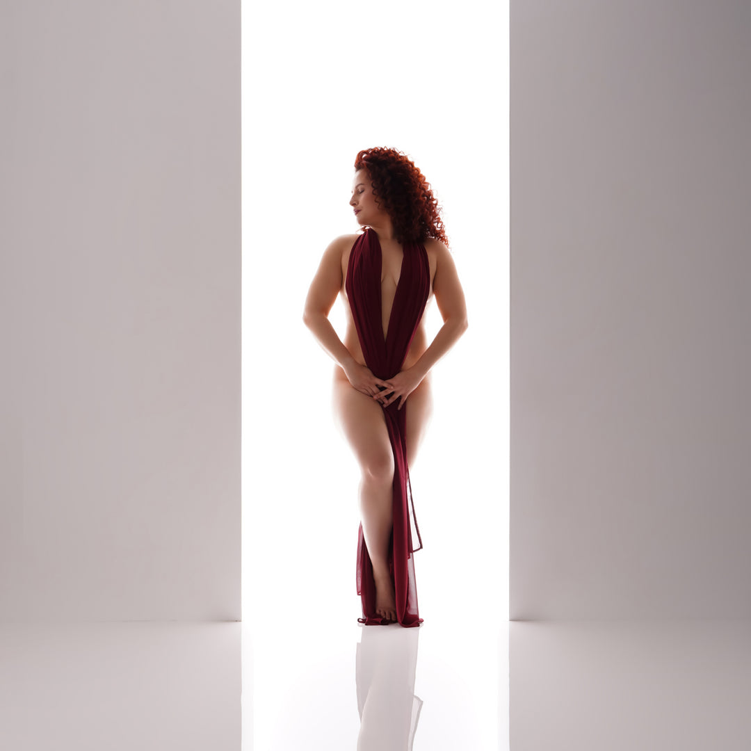 High key fine art portrait of a woman with red curls in burgundy chiffon standing in a bright doorway