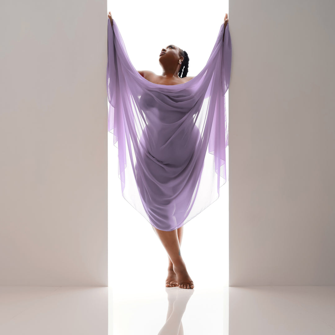 Woman draped in flowing lilac fabric poses in a bright backlit studio doorway for an elegant boudoir portrait.