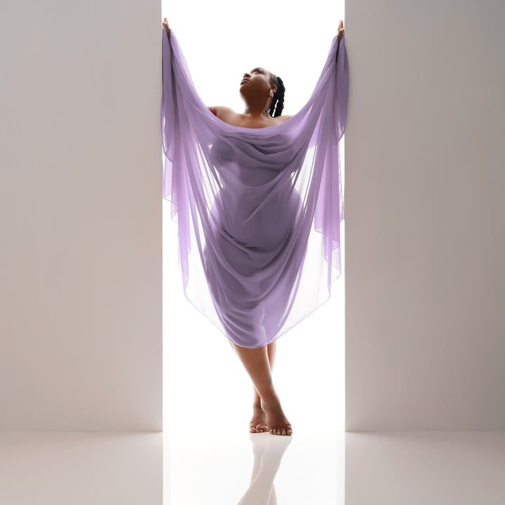 Woman draped in flowing lilac fabric poses in a bright backlit studio doorway for an elegant boudoir portrait.