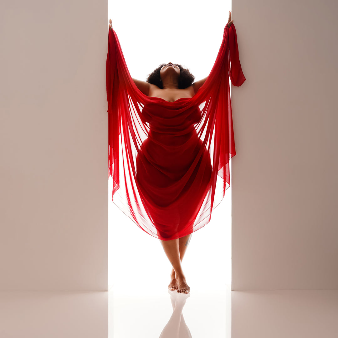 Retrato artístico de estilo «boudoir» de una mujer vestida con una vaporosa gasa roja, con los brazos elegantemente extendidos en el umbral de un luminoso estudio. 