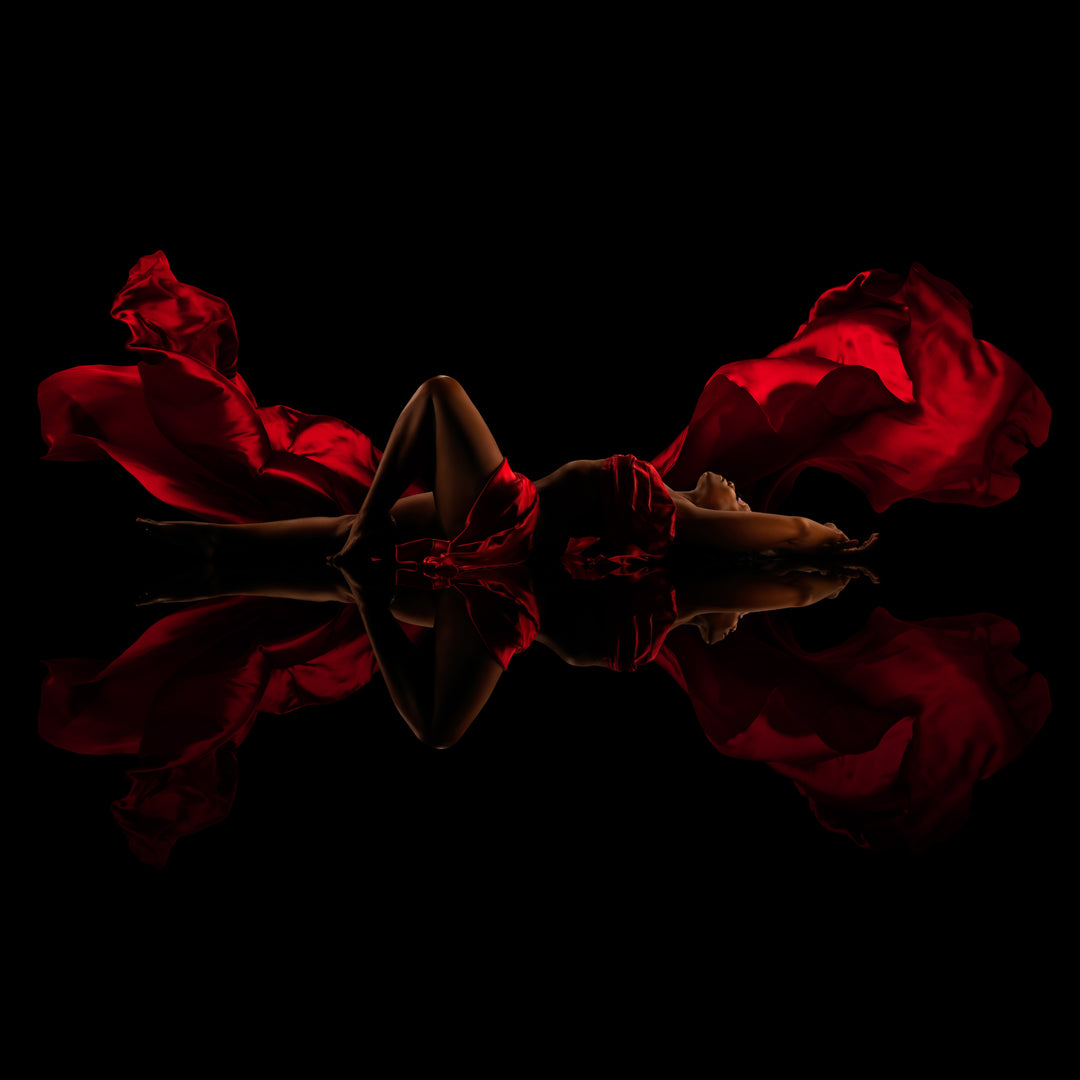 Woman reclines among flowing red fabric on a glossy black reflective floor for a dramatic fine art boudoir portrait.