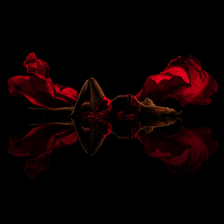 Woman reclines among flowing red fabric on a glossy black reflective floor for a dramatic fine art boudoir portrait.