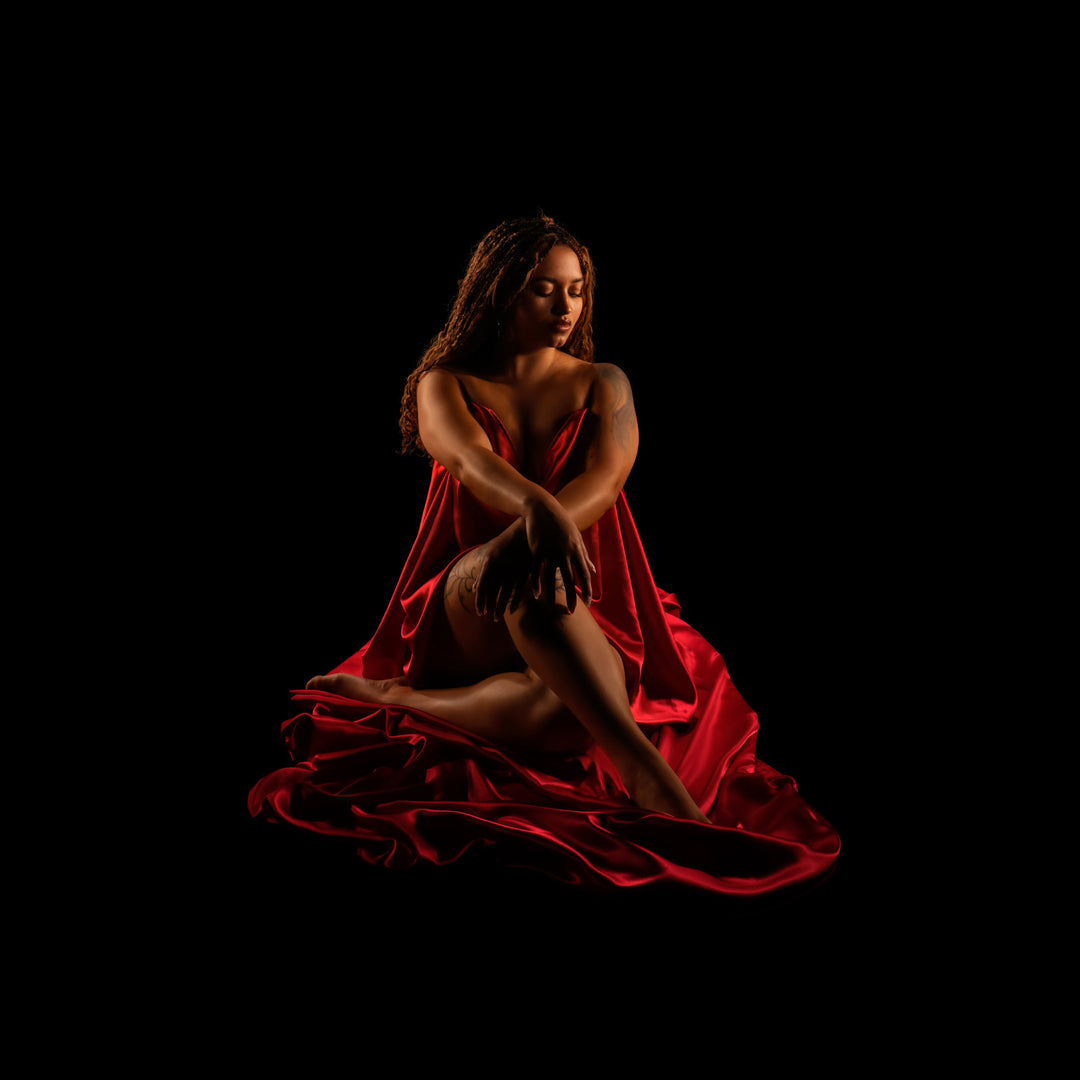 Woman seated and draped in rich red fabric poses against a black background for a dramatic studio boudoir portrait.
