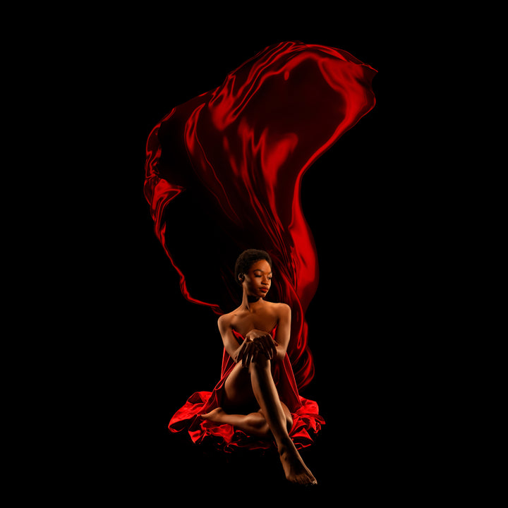 Fine art boudoir portrait of a woman seated as red satin billows overhead against a black backdrop