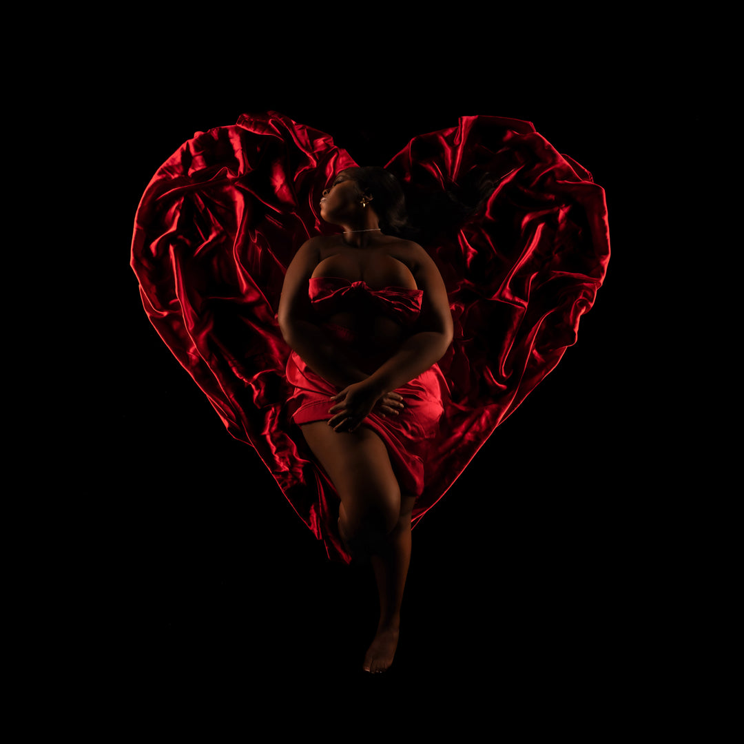 Fine art boudoir portrait of a woman in red satin arranged into a heart shape against a black backdrop